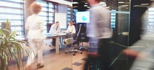 Stock picture of an office with a man and woman at a desk and two blurred moving figures of women