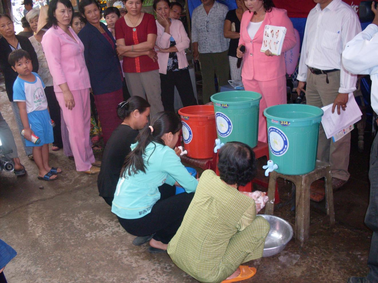 People crouching washing hands from buckets with taps and onlookers surrounding