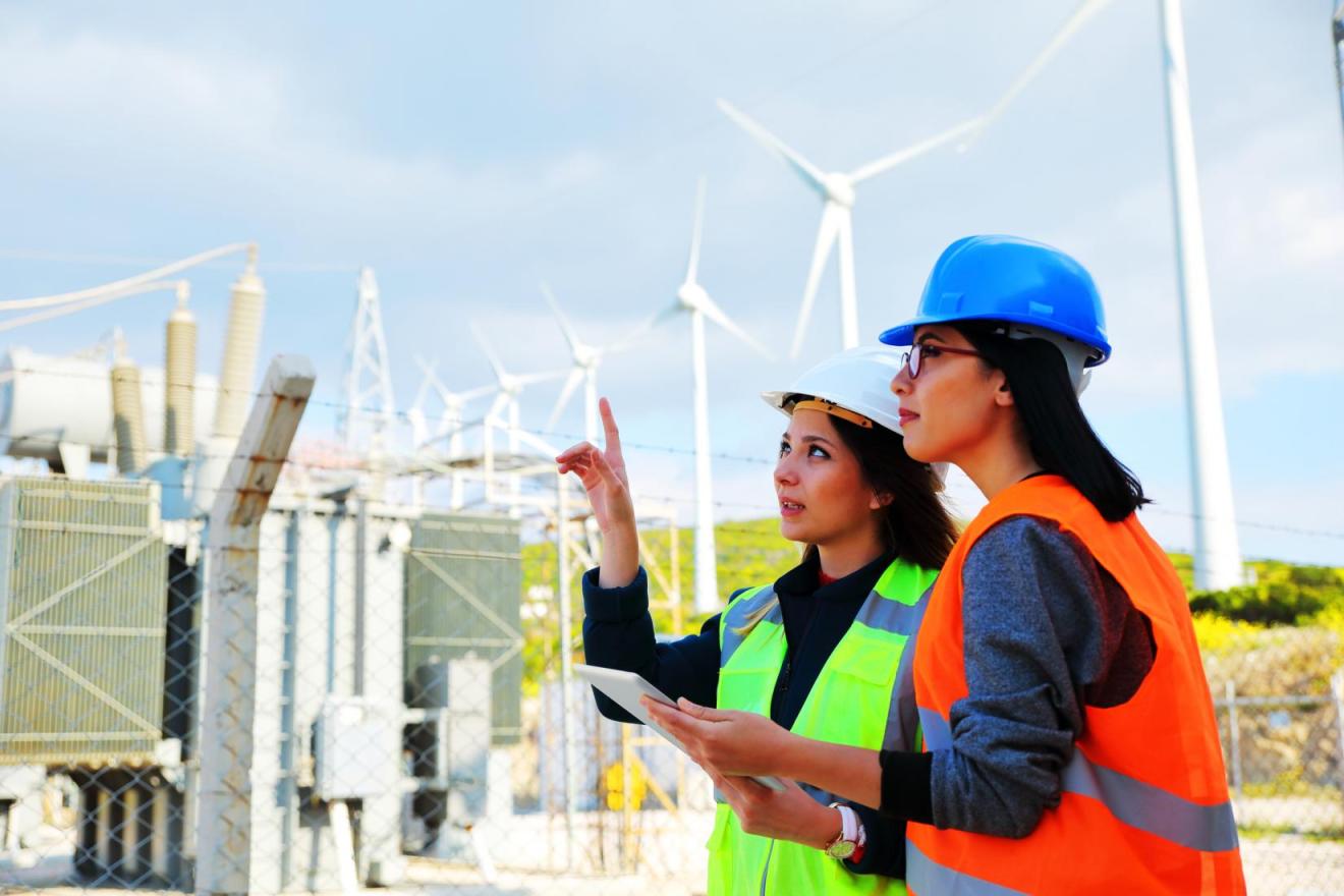 Two women in hi-vis gear stand in front of wind turbines. 