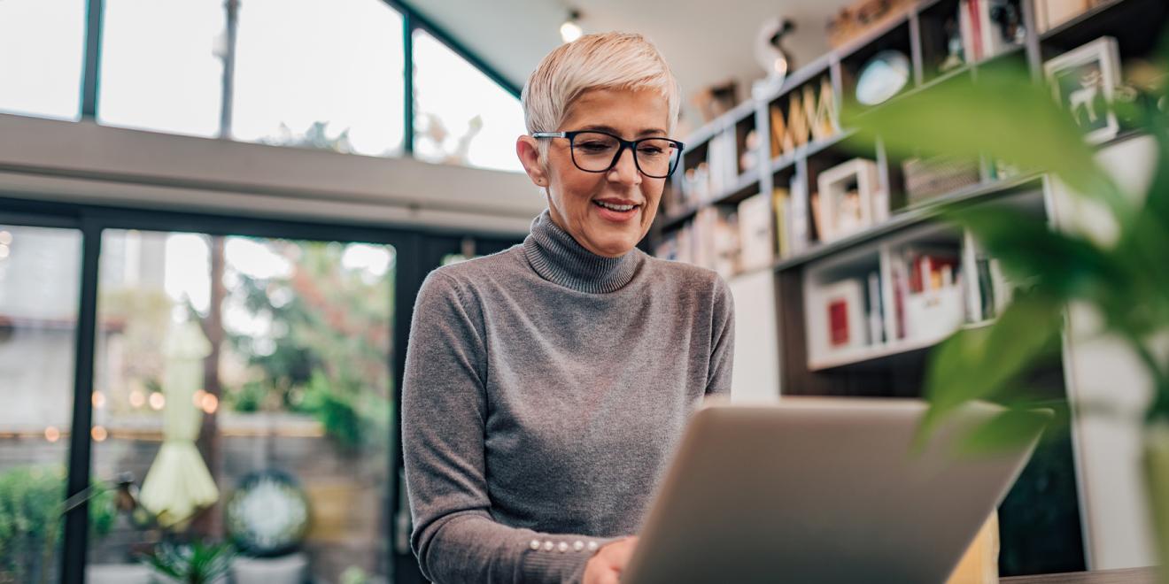 Woman with short blonde hair and glasses sitting at a table looking at a silver laptop 