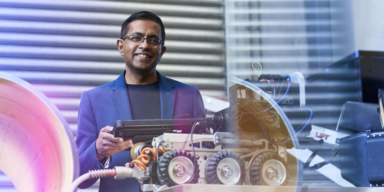 Man in a blue suit looking at the camera and holding a remote with a small robotic vehicle in front of him