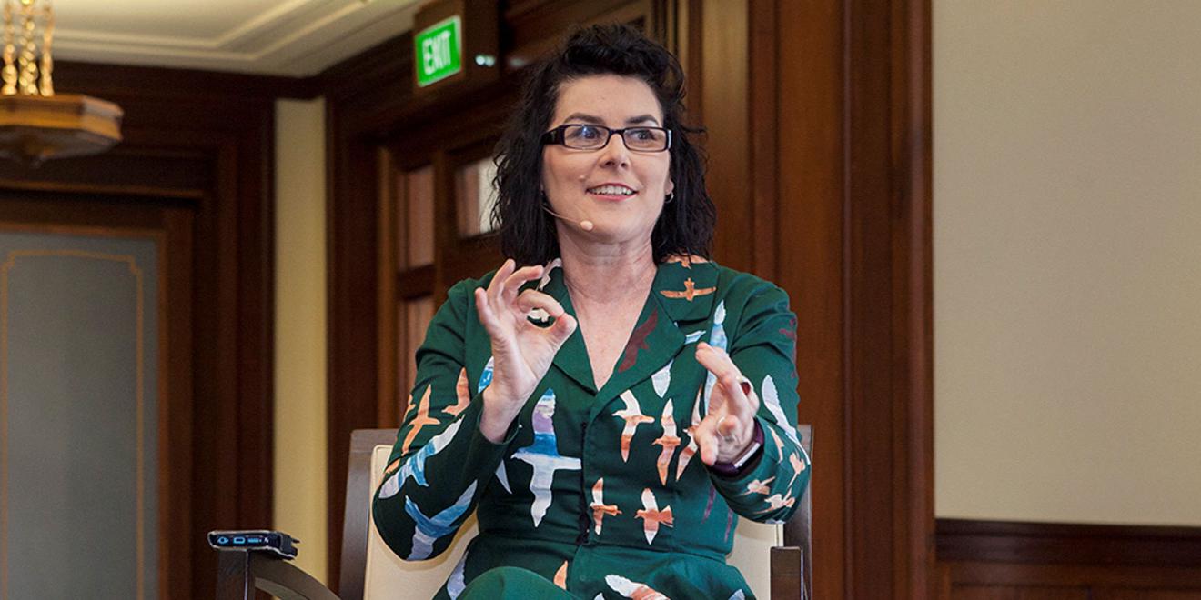 Photo of a woman with dark coloured hair and glasses seated in a green dress with colourful birds