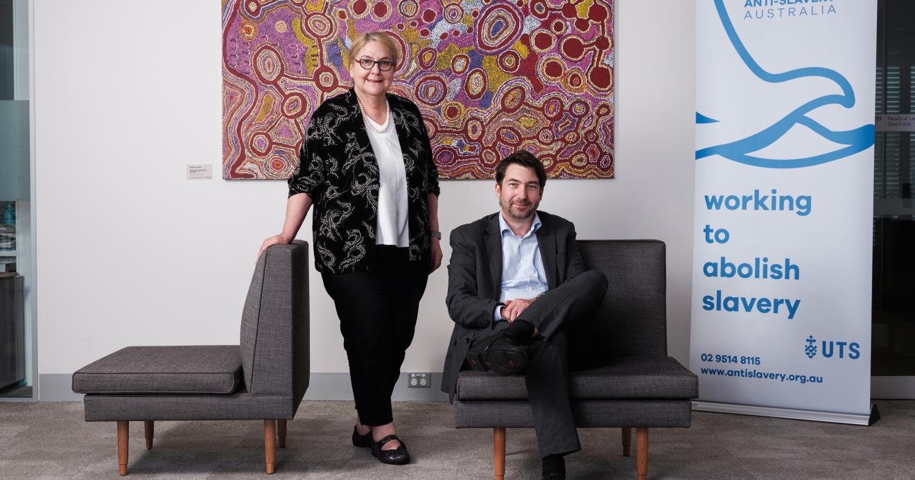 Woman standing next to a seated man in front of an indigenous artwork and a pull up banner for Anti Slavery Australia