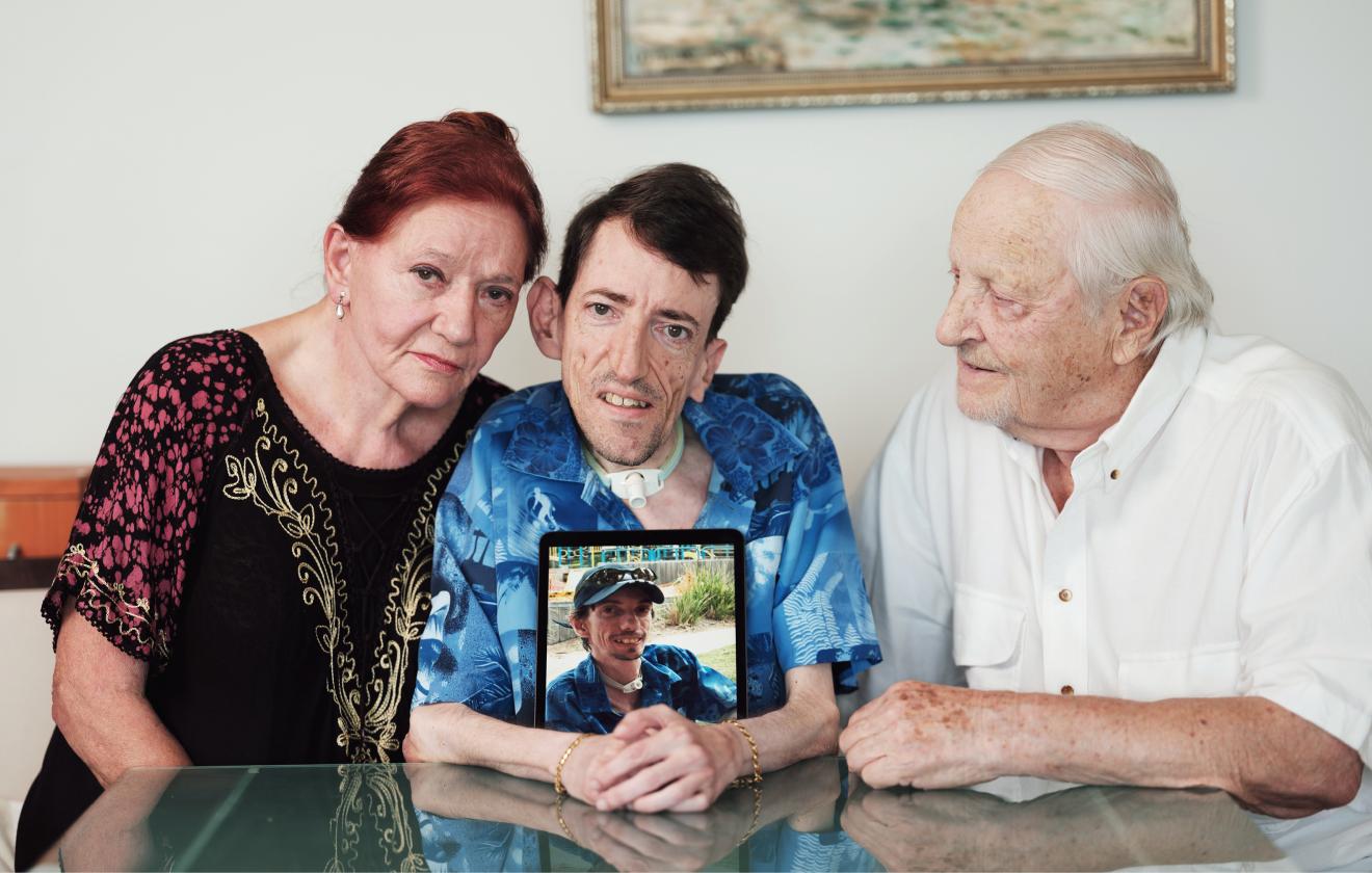 Mum, son and father seated at a table. Mum and son looking at the camera and father looking at the son. Son is holding a photo of his brother Lawrence.