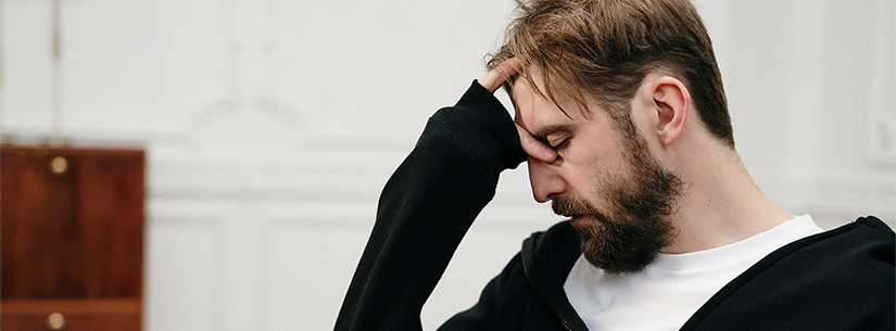 A man with short hair and a beard sits with his head resting on his hand. He is wearing a white t-shirt under a black jumper.
