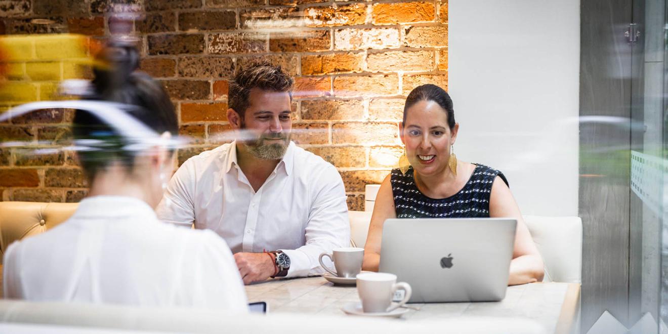 Three people sitting at a table behind glass, working on a laptop