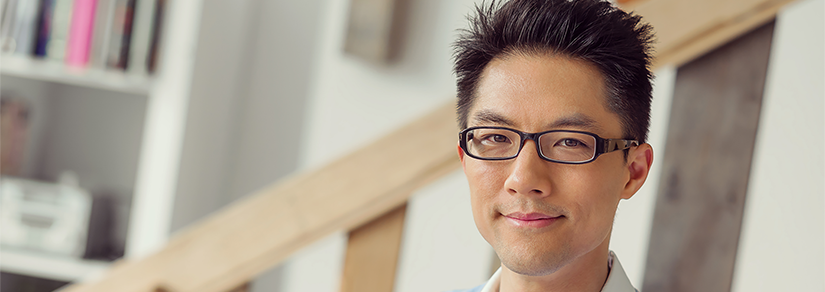 Young Asian man in glasses, white collared shirt and blue jumper sitting at a desk with a white laptop