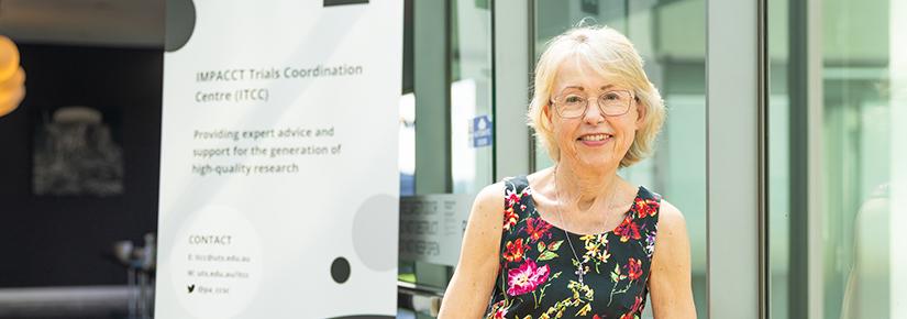 Imelda Gilmore has short blonde hair and is wearing sleeveless top that is black with a flora print. She wears glasses and is smiling at the camera.