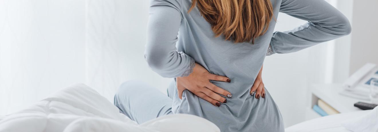 a person sits on a bed and clutches their back, seemingly in pain