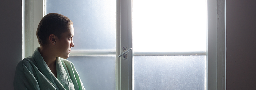 Young woman with short hair sits in front of window. She is wearing a bath robe and looking out the window, which has frosted glass. 