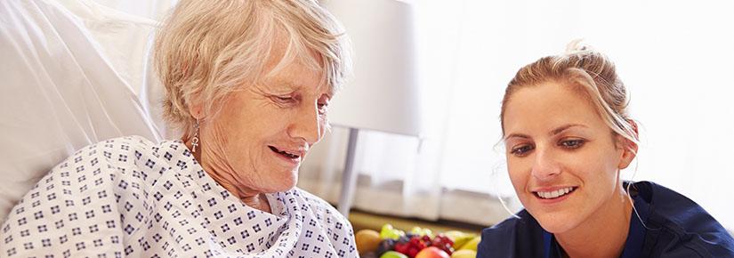 Young woman talking to elderly woman who is in bed looking at a tablet.