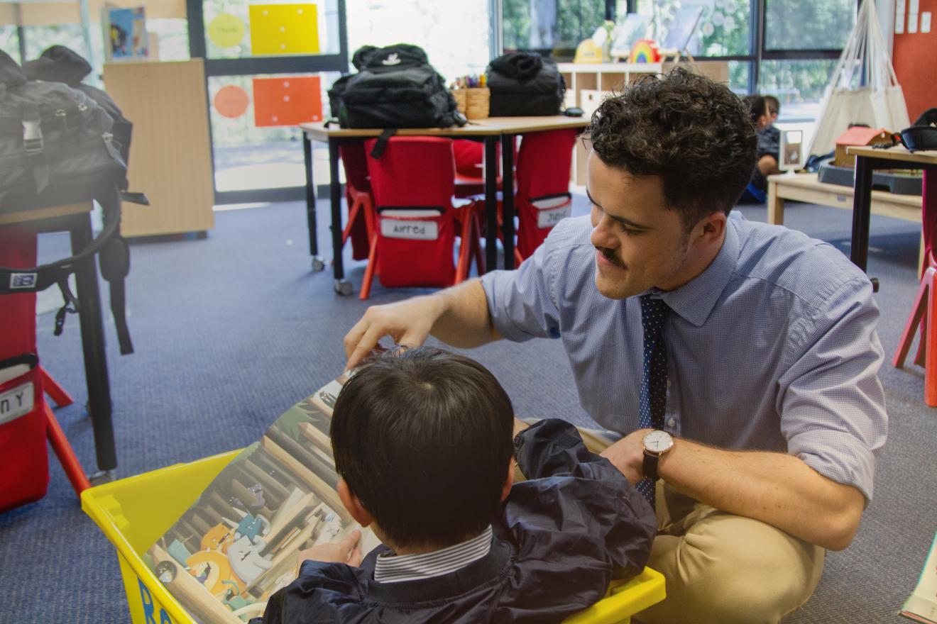 Tom crouches down to help one of his students reading a picture book