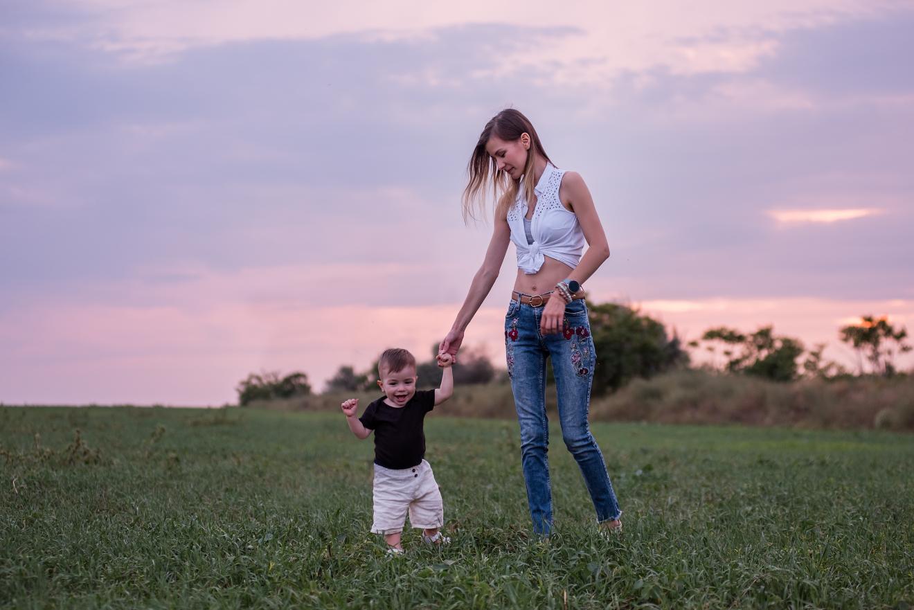 Little boy gleefully taking first steps in green field, guided by young mother, with soft sunset hues painting the sky. Woman and child walking in the field