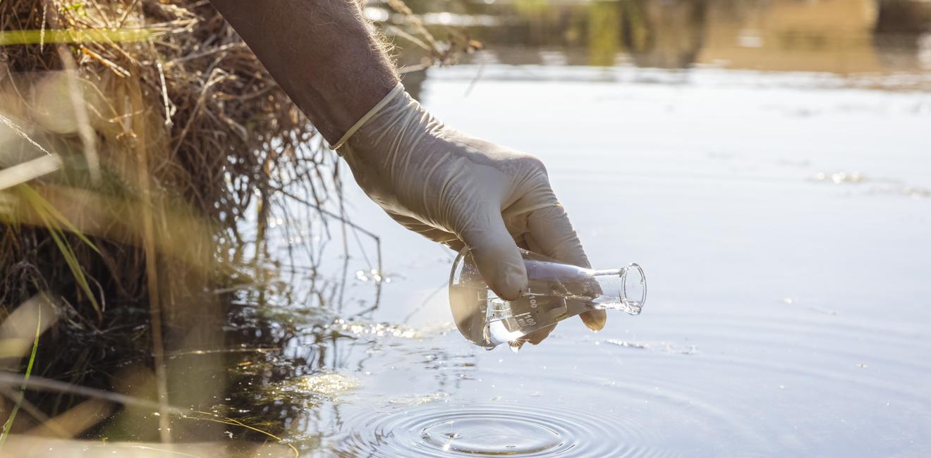 A gloved hand holding a beaker above a body of water