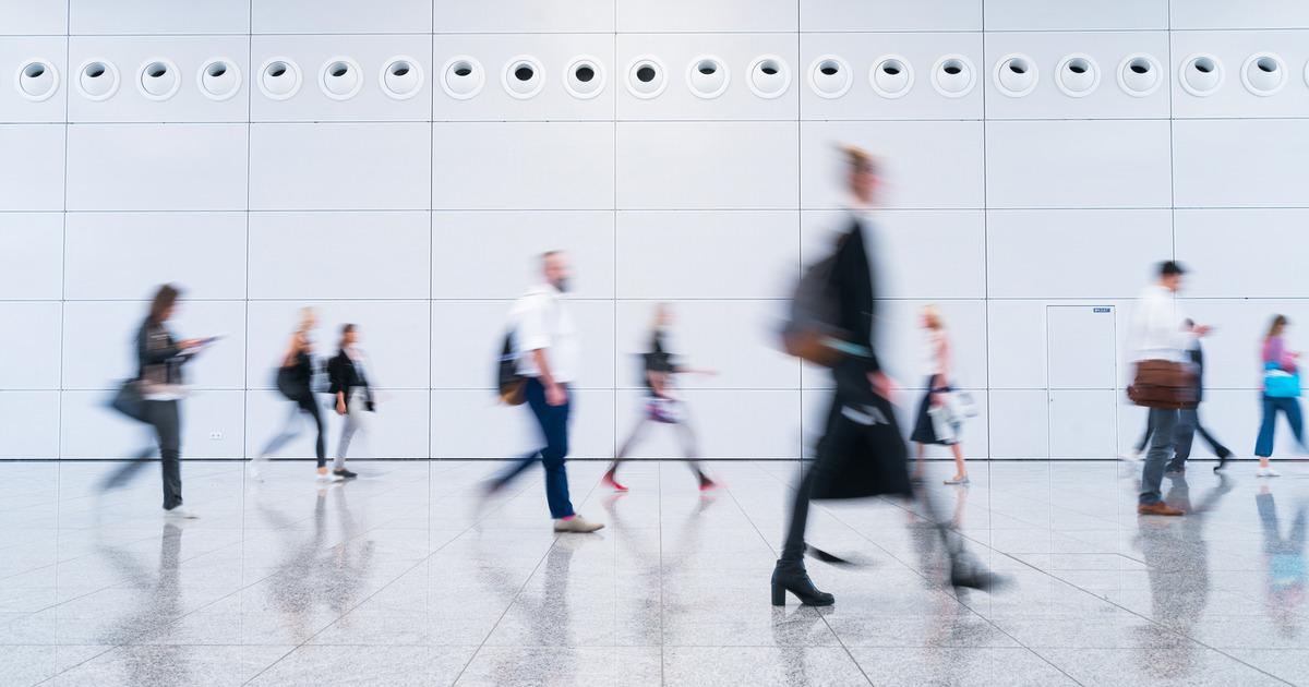 Wide shot of the lobby of a large white office building. People dressed in business clothing are walking past in a blur.