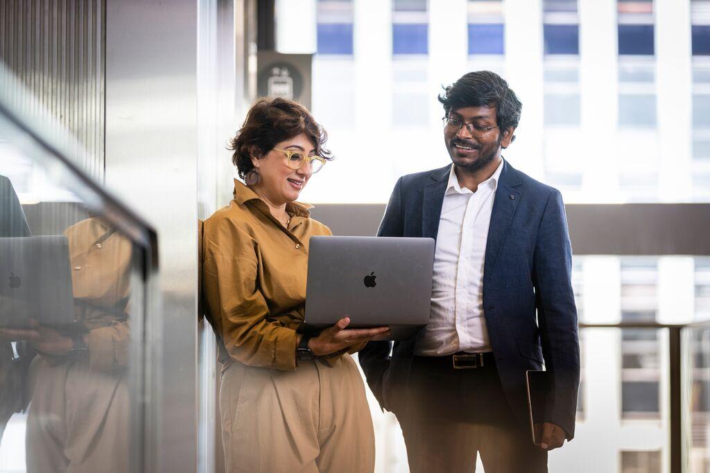 Woman leaning against a wall with a yellow top holding a laptop and a man in a suit looking at the laptop