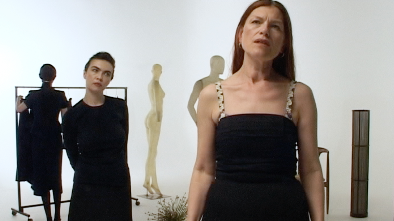 Two women in black dresses stand in a minimalist gallery space with mannequins and clothing racks behind them, creating a fashion-focused, contemplative scene.