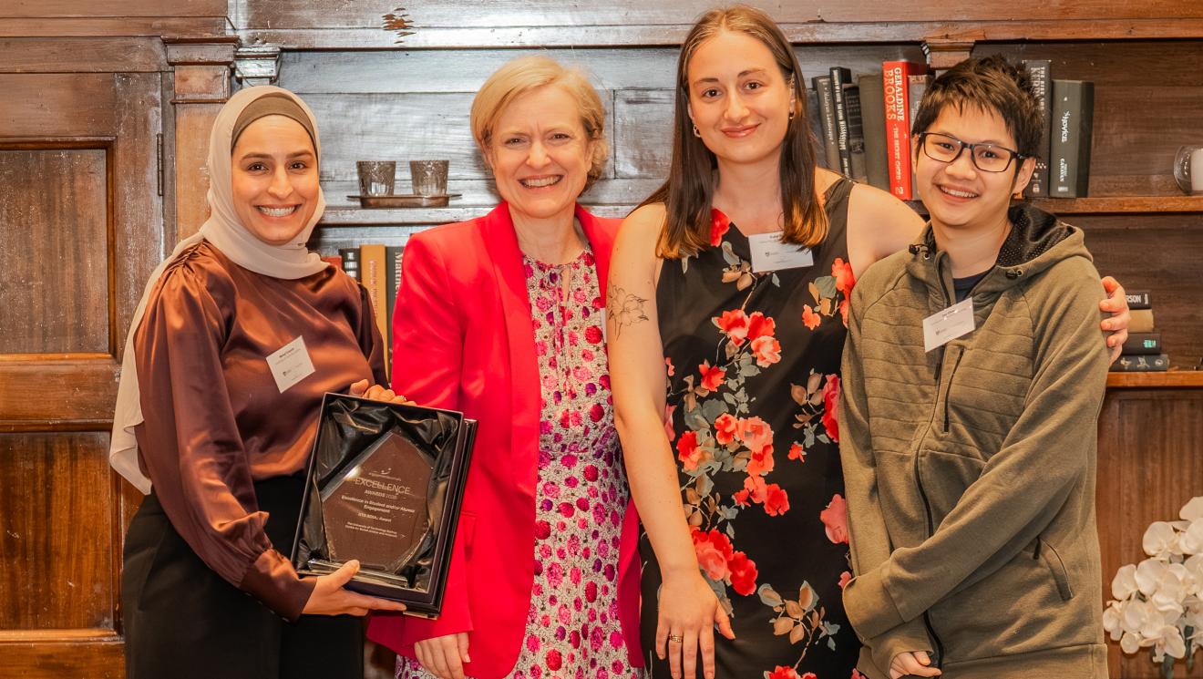 Four people stand closely together, smiling warmly. One holds an award plaque. They stand in front of wooden shelves filled with books, creating a celebratory atmosphere.