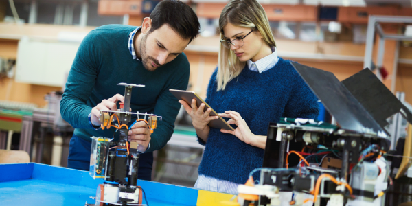 A male and female collaborate on an electronic engineering project over a bench