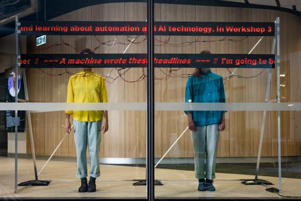 Two people, one in a blue shirt and one in a yellow shirt, stand with their faces obscured by a digital sign with red text on it. 
