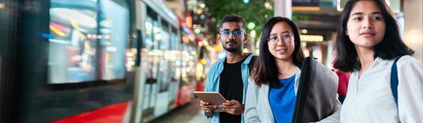 Three international students waiting on a platform as a light rail passes