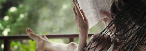 A person sits on a porch with her feet up and crossed while reading a book.