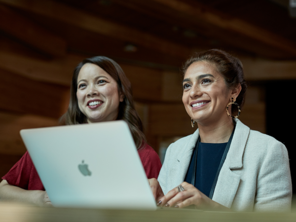 Two UTS female staff members smiling while looking off camera. One is typing on a Apple Mac laptop.