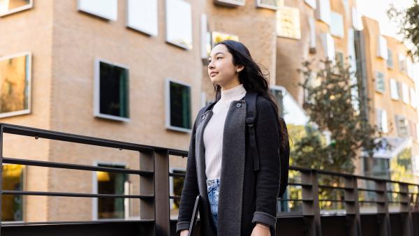 A young female student with a contemplative expression walking past university buildings, carrying a backpack