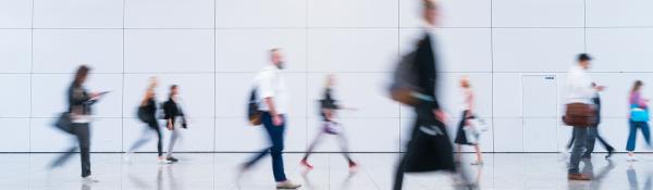 Wide shot of the lobby of a large white office building. People dressed in business clothing are walking past in a blur.