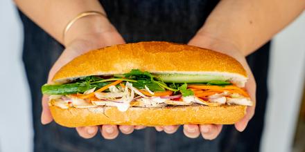 Hands hold a white breadroll full of fresh salad, meat and sauce