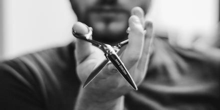 A man with a well-trimmed beard holds a pair of sharp scissors towards the camera