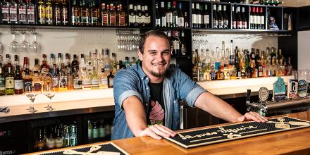 A barman in a denim shirt standing behind a bar 