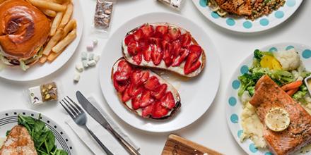 Plates of brightly coloured food cover a table, with means including fish, salads, pizza, a burger and chips, and strawberry and ricotta on toast.