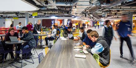Students sit at tables eating and looking at computers in a busy food court