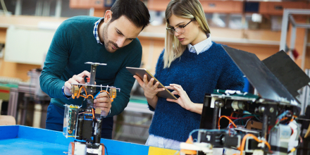 A male and female collaborate on an electronic engineering project over a bench