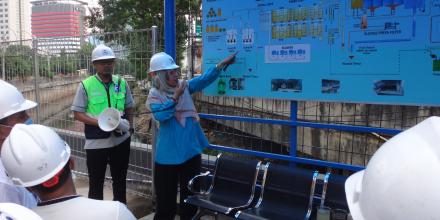 A woman in a hard hat pointing at a blue sign next to a man in a green high-visibility vest.