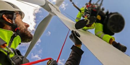 An energy worker in yellow hi-vis gear stands at the base of a wind turbine. He is looking up at another worker who is on scaffolding.