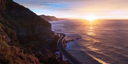 Windy road with mountains on the left and the sea to the right. The sun is setting in the background.