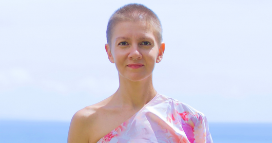 Christina Giarmatzi standing in front of the ocean and wearing a floral off-the-shoulder dress.