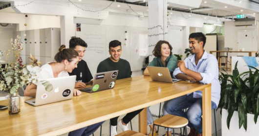 Five young people smiling as they sit around a table in front of their laptops