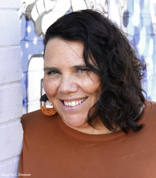 headshot image of woman smiling at camera. she has brown curly hair and orange/brown earrings and top