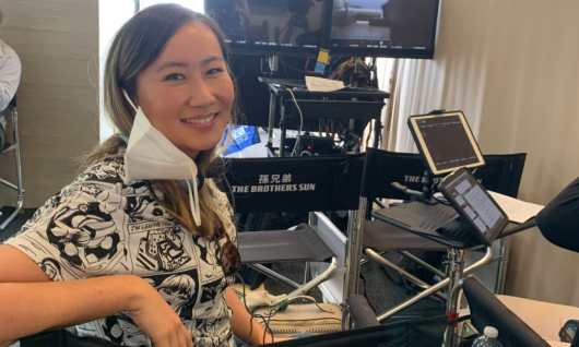 image of woman sitting in Director's chair on film set and turning around to smile at camera. There is a film camera behind her.