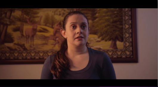 a young woman looking at something off camera. there is a painting behind her. she is inside somewhere. she has long brown hair in a low ponytail. sh