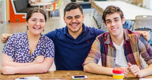 Three students sitting at a desk with papers and a coffee cup, smiling.