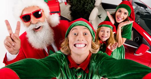 A man takes a selfie with Santa with a woman and young girl in the background.