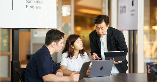 Two students looking at Apple laptops while a course instructor points to the screen.