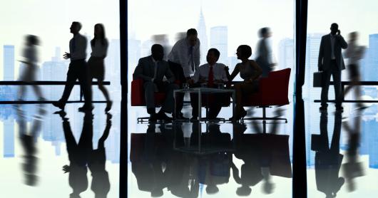 a group of people gathered in a large office around a table with large windows in the background.