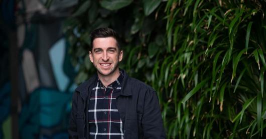 A man stands smiling at the camera, a green wall in the background