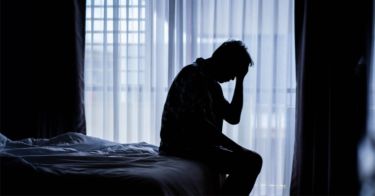 Silhouette of a man sitting on a bed with his hand held to his head. Filtered light is coming through the window in the background, which has net curtains.