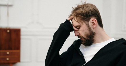 Male patient sits in a chair with his hand held to his head. He has a beard and short hair. He is wearing a white tee shirt with a dark coloured jacket.